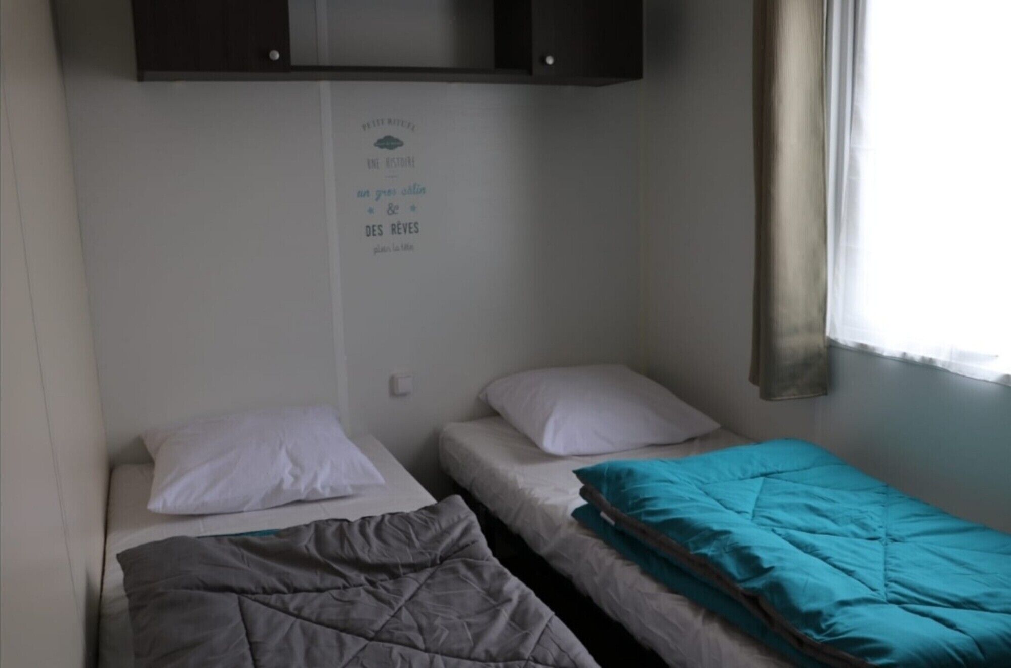 Quarto de dormitório com duas camas individuais, uma prateleira fixada na parede e uma janela com cortinas.