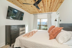 A bedroom with a bed, a TV mounted on the wall, a ceiling fan, and a window with curtains.