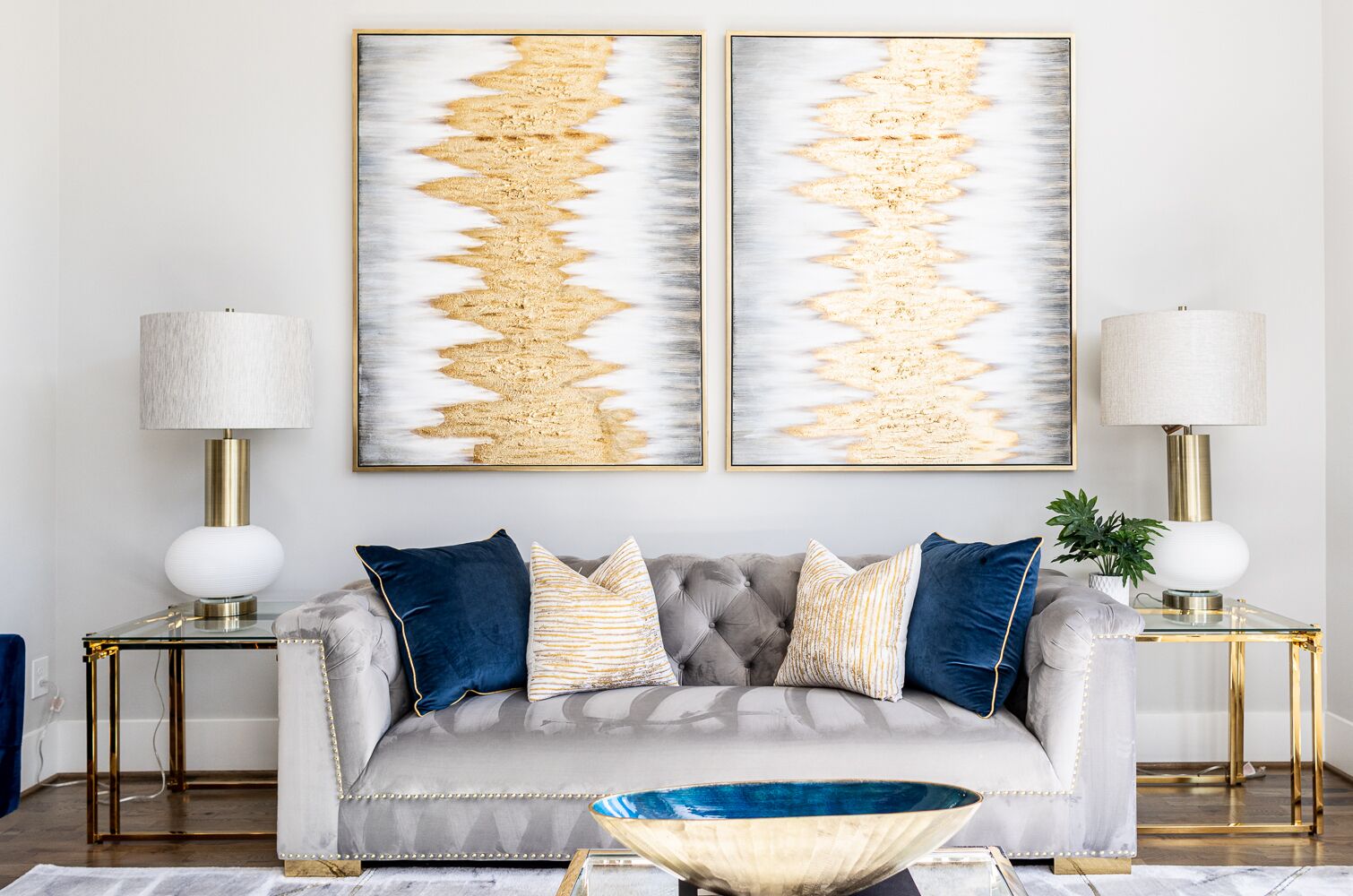 Een woonkamer met een grijze gestofte bank, twee bijzettafels met lampen, en twee grote abstracte schilderijen aan de muur.