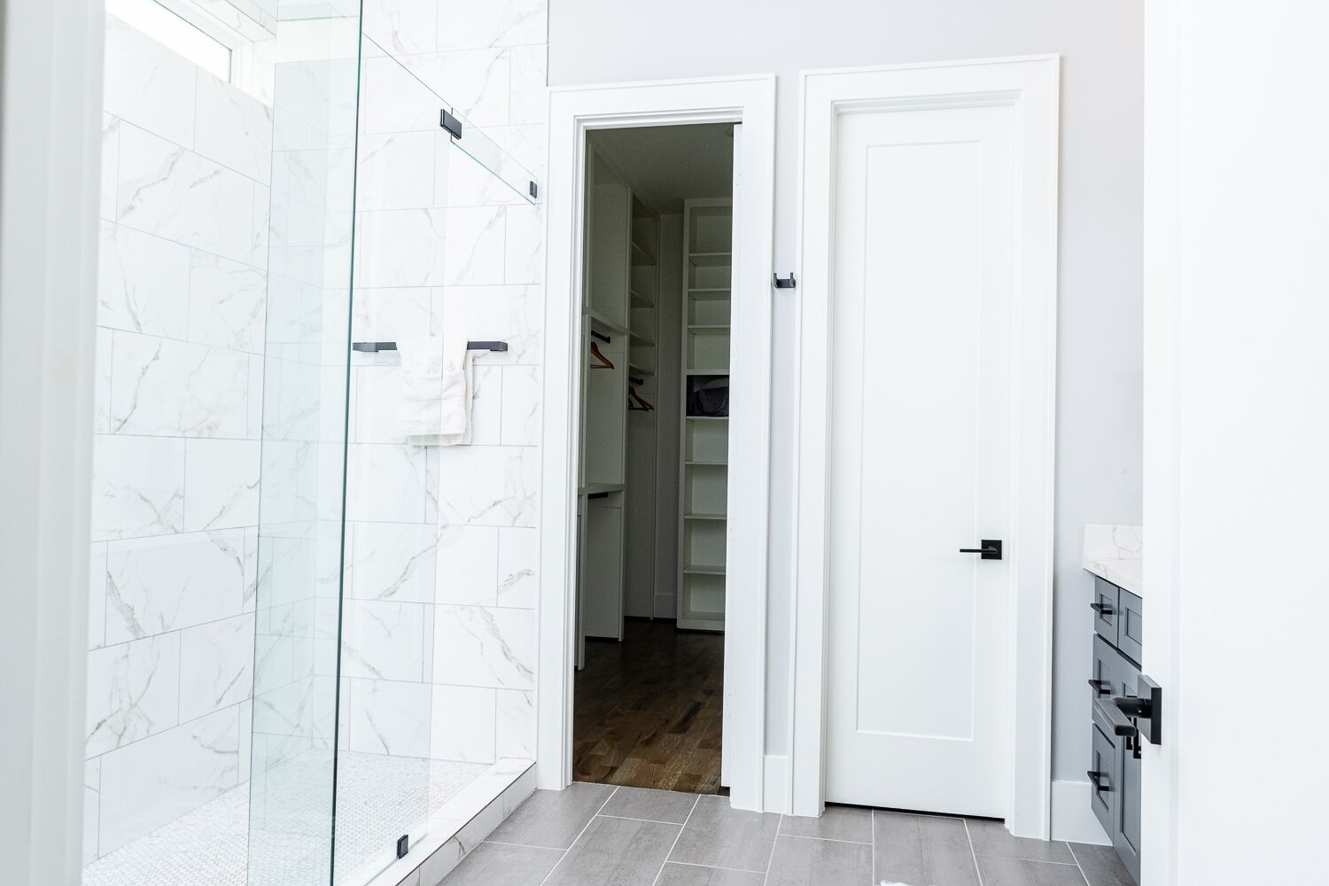 A modern bathroom with white marble tiles, a glass shower enclosure, and a white door leading to a hallway with a wooden floor.