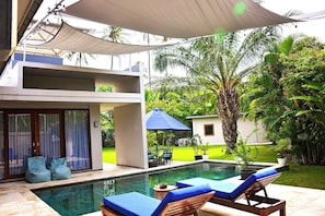 A swimming pool with lounge chairs, a covered patio, and a modern building with a glass door.