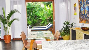 A modern dining area with a glass table, cushioned chairs, and a view of lush greenery outside.