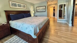 A bedroom with a large bed, wooden headboard, and a ceiling fan. There are framed pictures on the wall and a hallway leading to a closet.
