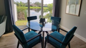 A dining area with a glass table and four chairs, large windows with curtains, and a view of a lake and greenery.