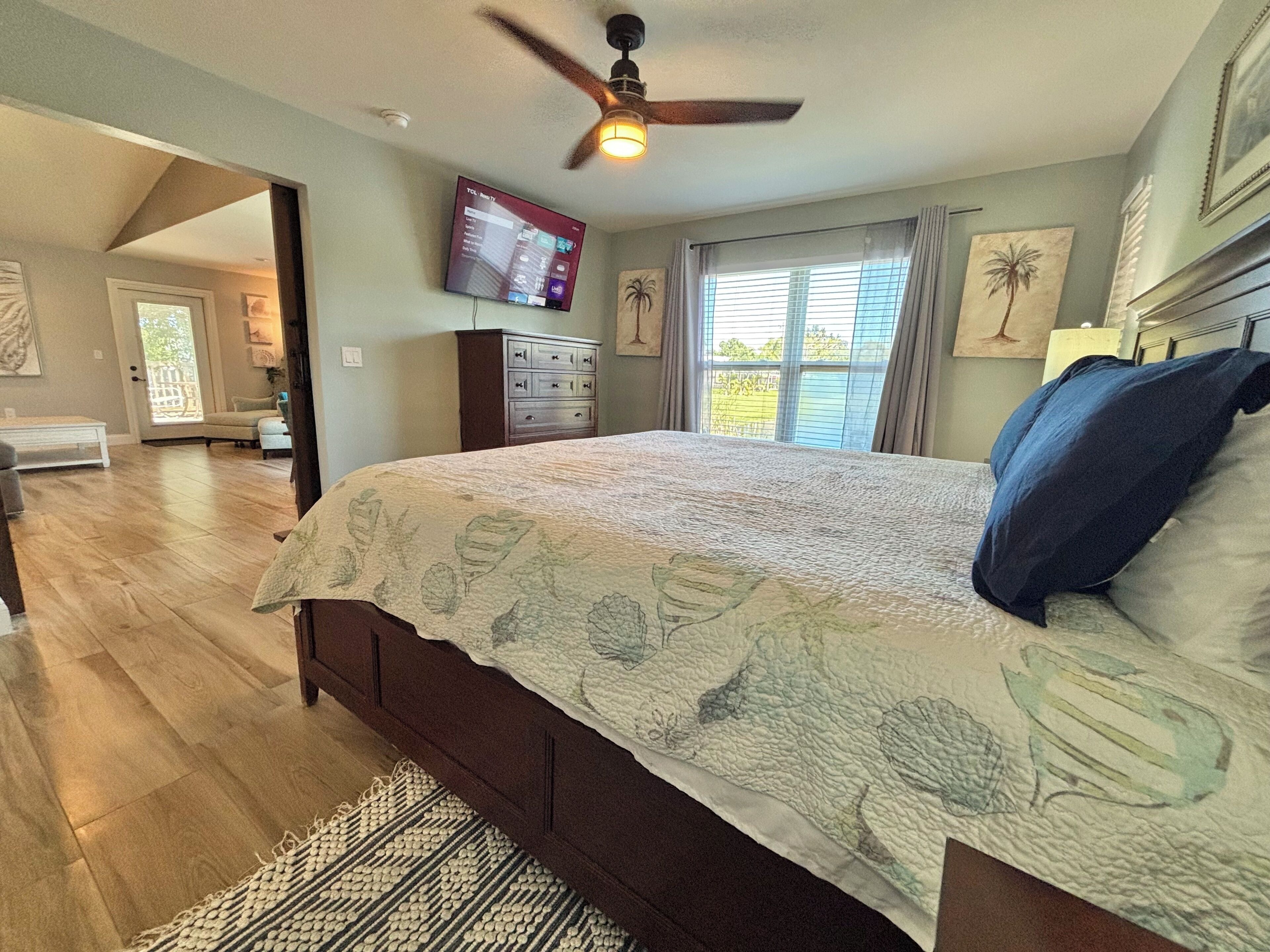 A bedroom with a large bed, wooden flooring, a ceiling fan, a TV mounted on the wall, and a window with curtains.