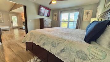 A bedroom with a large bed, wooden flooring, a ceiling fan, a TV mounted on the wall, and a window with curtains.