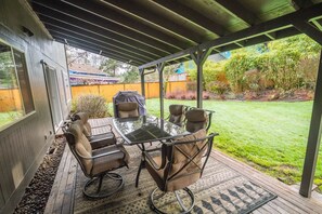 An outdoor patio with a glass table and four chairs, a covered roof, and a view of a backyard with a lawn and trees. - 6BR-Spacious-Bright-Modern (Eugene)