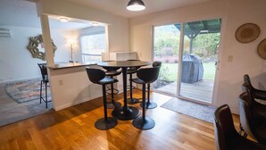 A modern kitchen with a dining area, featuring a round table and four chairs, a pendant light, and a view of the backyard through a sliding glass door. - 6BR-Spacious-Bright-Modern (Eugene)
