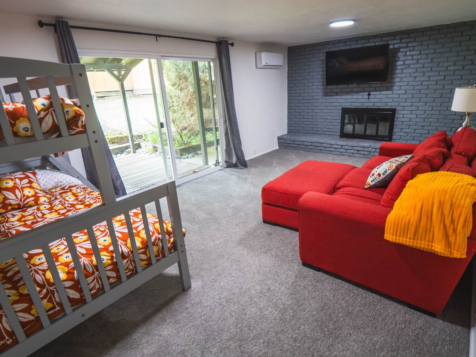 Una habitación con una cama litera, un sofá rojo, una chimenea y un televisor montado sobre una pared de ladrillo.
