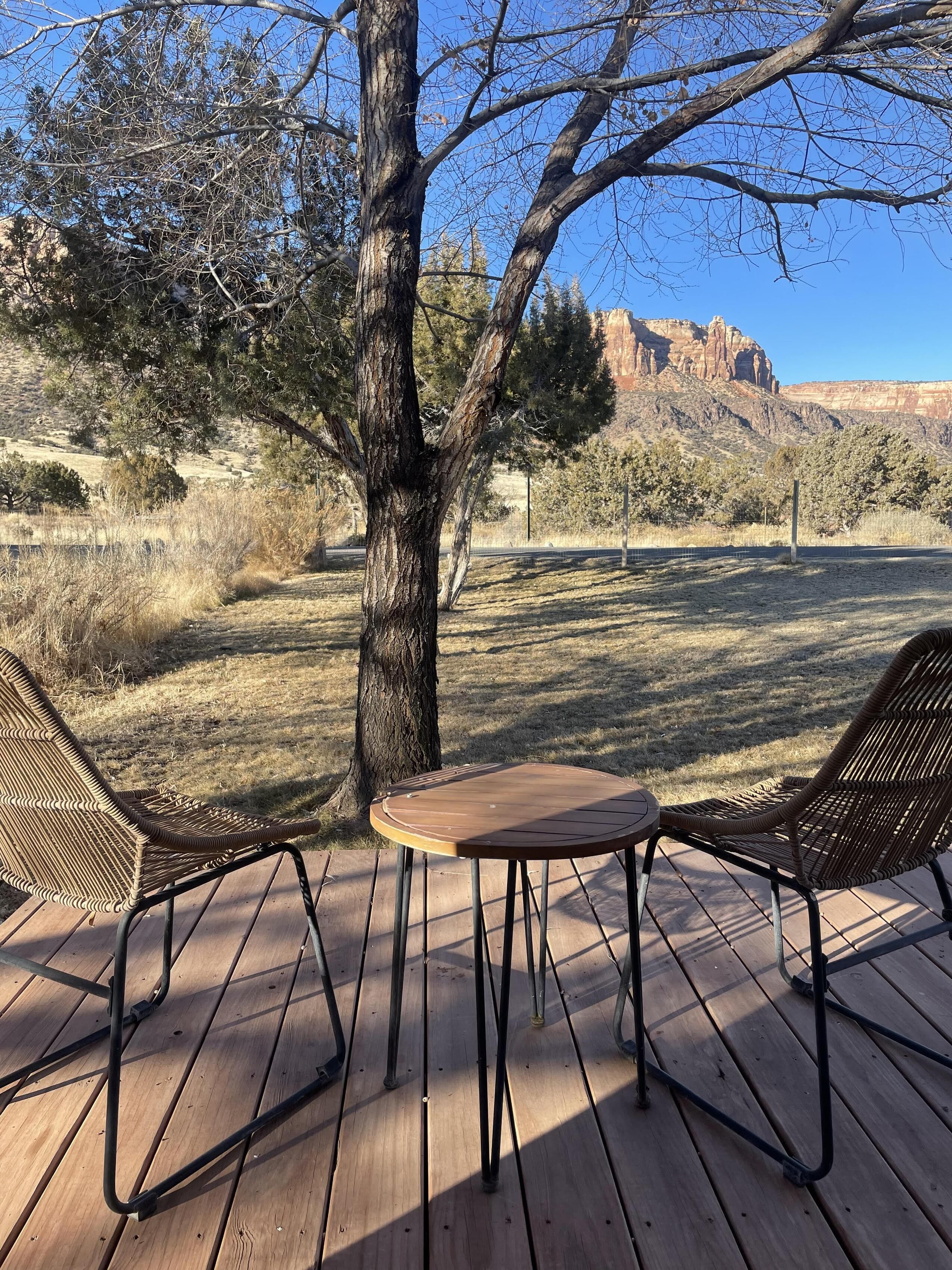 A wooden deck with two wicker chairs and a small table, surrounded by dry grass and a large tree.