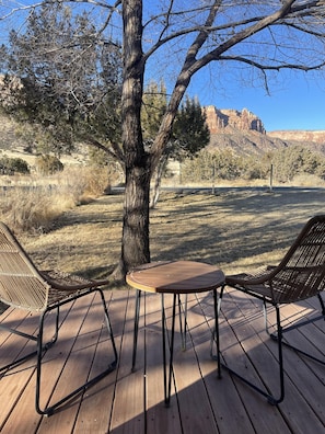 A wooden deck with two wicker chairs and a small table, surrounded by dry grass and a large tree. - Rabbitbrush: Trail Access at Your Door (Grand Junction)