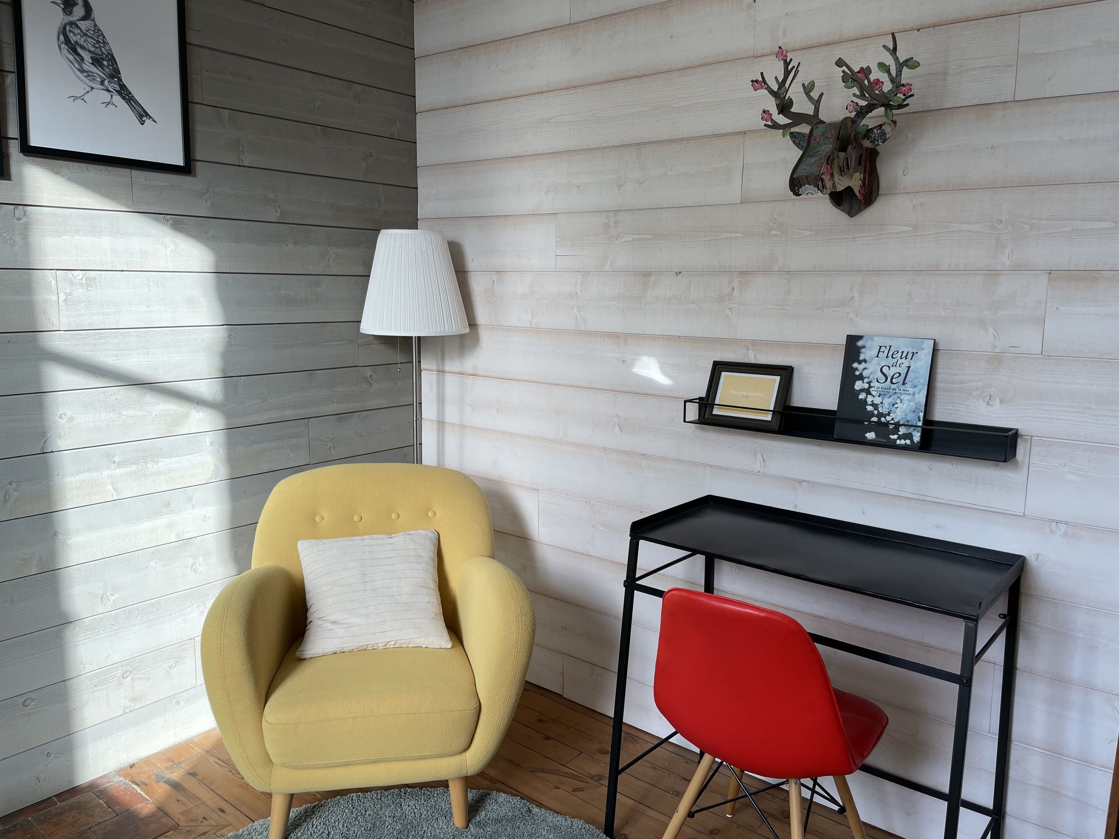 Ein Zimmer mit einem gelben Sessel, einem roten Stuhl, einem schwarzen Schreibtisch und einem an der Wand befestigten Regal mit Dekorationen und einem gerahmten Bild.
