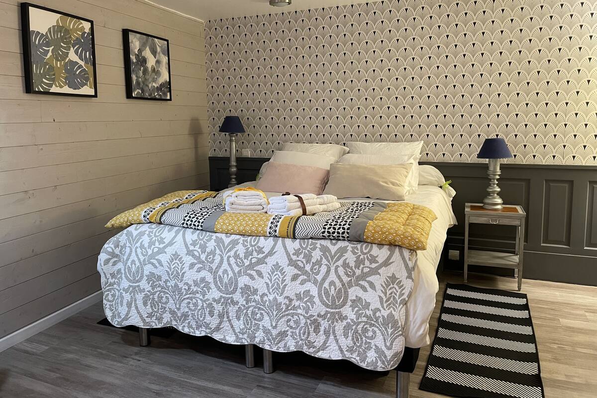 Een net opgemaakt bed met een bedspreide en kussens, een nachtkastje met een lamp, en een decoratieve wand met een herhalend patroon.