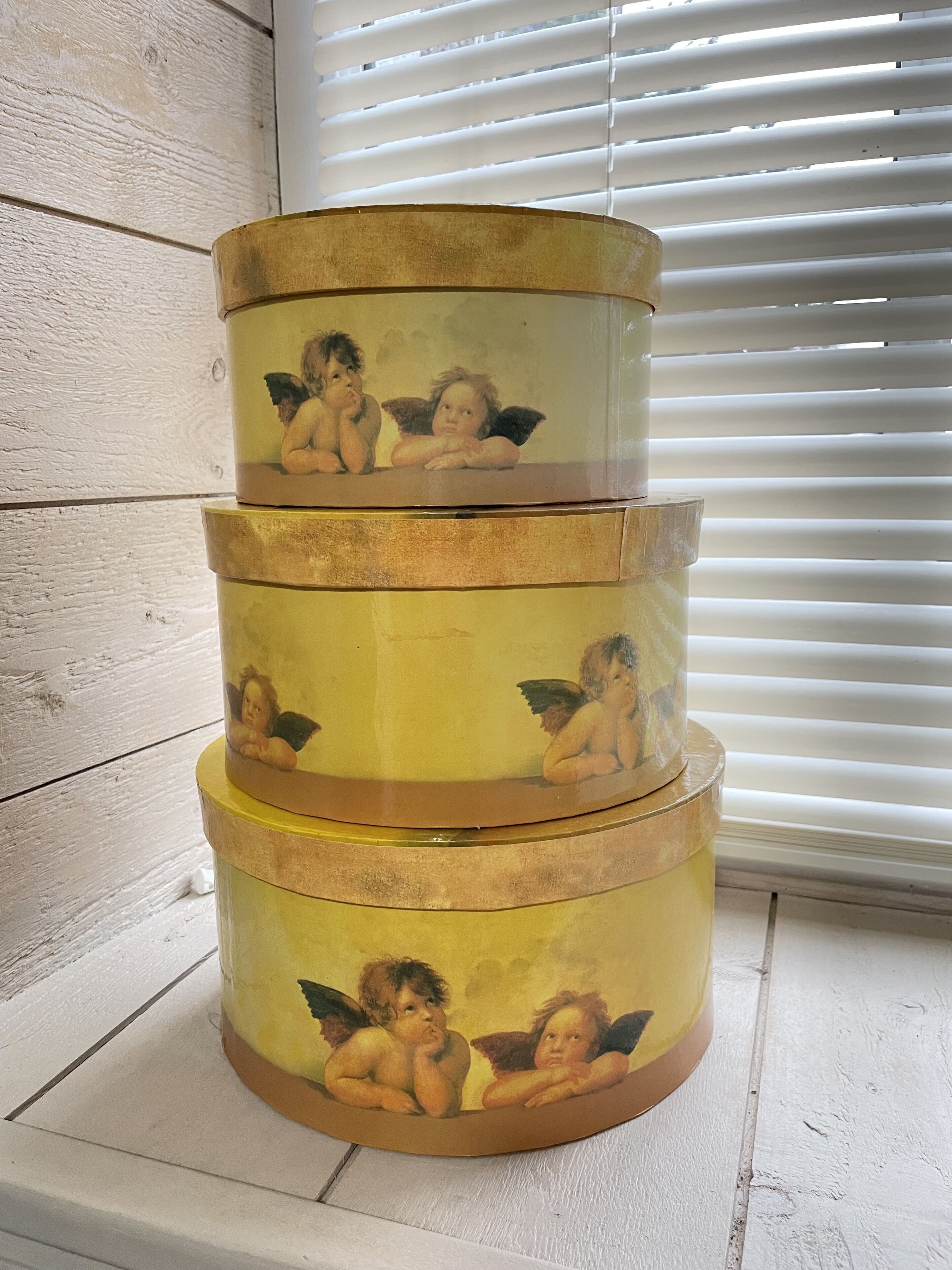 A three-tiered cylindrical container with angelic figures on each tier, placed against a white wooden wall with blinds in the background.
