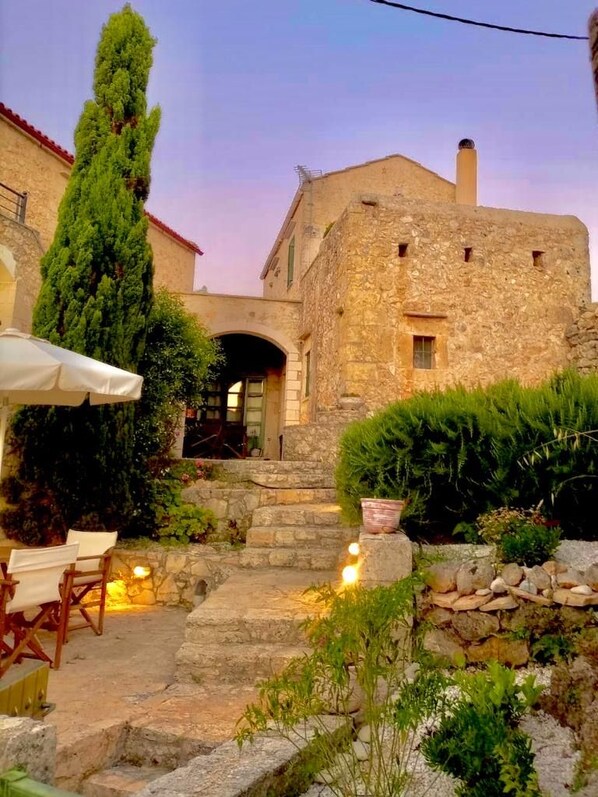 A stone building with a staircase leading to an entrance, surrounded by greenery and outdoor seating.