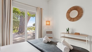 A hotel room with a bed, a view of the outdoors, a round mirror, a wall-mounted light, and a small shelf with a plant. - Villa 'Artemis' with Sea View, Wi-Fi, and Air Conditioning in Grimaud (Grimaud)