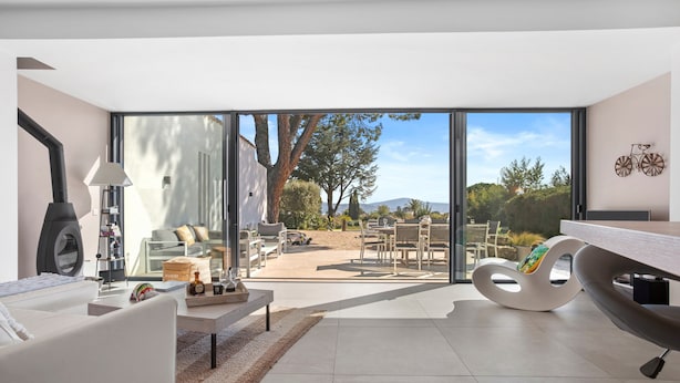 A modern living room with a sofa, coffee table, and a unique chair, a large window with a view of a garden and mountains, and a dining area with chairs and a table. - Villa 'Artemis' with Sea View, Wi-Fi, and Air Conditioning in Grimaud (Grimaud)