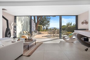 A modern living room with a sofa, coffee table, and a unique chair, a large window with a view of a garden and mountains, and a dining area with chairs and a table. - Villa 'Artemis' with Sea View, Wi-Fi, and Air Conditioning in Grimaud (Grimaud)