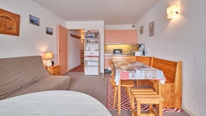 Ein kleiner Raum mit einem Bett, einem Esstisch mit Stühlen, einer Küchenzeile und einer Tür zu einem weiteren Raum.