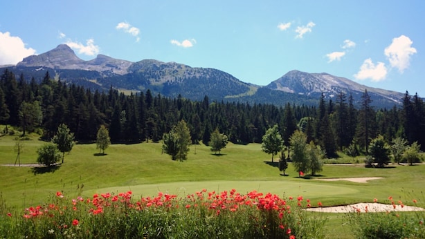Ein Golfplatz mit Bergblick, Bäumen und roten Blüten im Vordergrund.