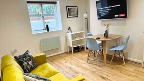 A modern living room with a yellow sofa, a dining table with chairs, a flat-screen TV mounted on the wall, and a window with a view of greenery outside. - Charming Period Apartment, Near Uni & Hospital, Parking (Reading)