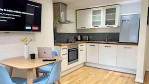 A modern kitchen with white cabinets, a stainless steel range hood, and a built-in oven. There's a dining area with a wooden table and blue chairs, and a flat-screen TV mounted on the wall. - Charming Period Apartment, Near Uni & Hospital, Parking (Reading)
