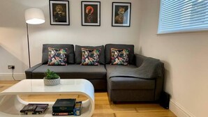 A modern living room with a grey sofa, colorful cushions, a white coffee table with books and a plant, and three framed artworks on the wall. - Period Apartment for 3, Near Uni & Hospital, Parking (Reading)