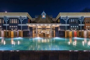 A hotel with a large pool in the foreground, a central entrance with a roof, and multiple windows on the facade.