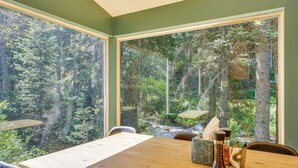 A dining table with chairs, a wooden table, and a view of a forest through large windows. - Solitude Mountain Cabin: Creek-Side View & Hot Tub (Brighton)