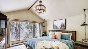 A bedroom with a large window, a bed with a blue bedspread, a wooden headboard, and a ceiling fan. - Solitude Mountain Cabin: Creek-Side View & Hot Tub (Brighton)