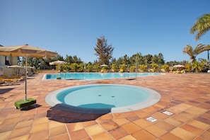 Une piscine entourée de palmiers, avec des chaises longues et des parasols, sous un ciel dégagé.
