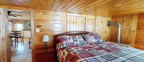 Una habitación con paneles de madera, una cama, una mesita de noche, una lámpara y una puerta que da a otra habitación.