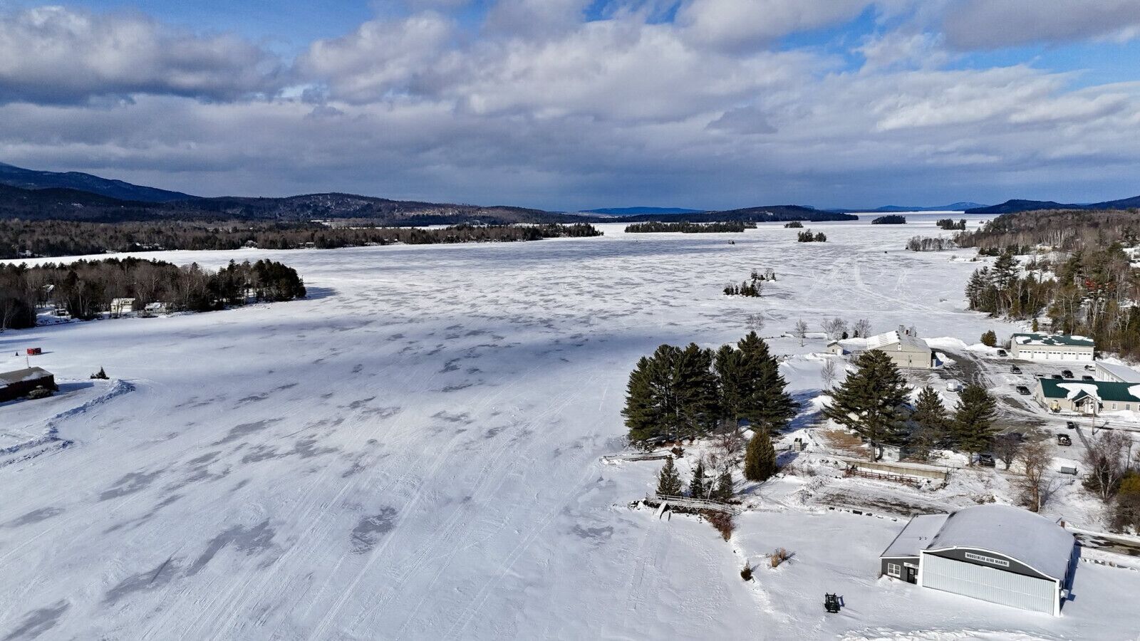 A snow-covered landscape with a small building complex, including a large white structure with a green roof, surrounded by trees and open fields.