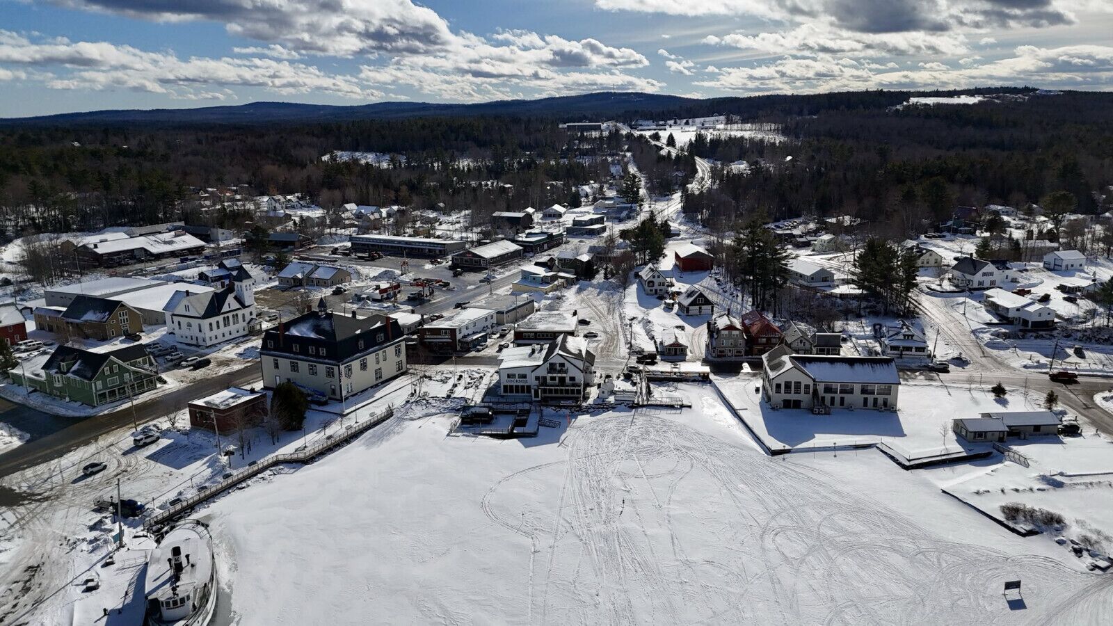 An aerial view of a small town with snow-covered streets and buildings, surrounded by forests and mountains.