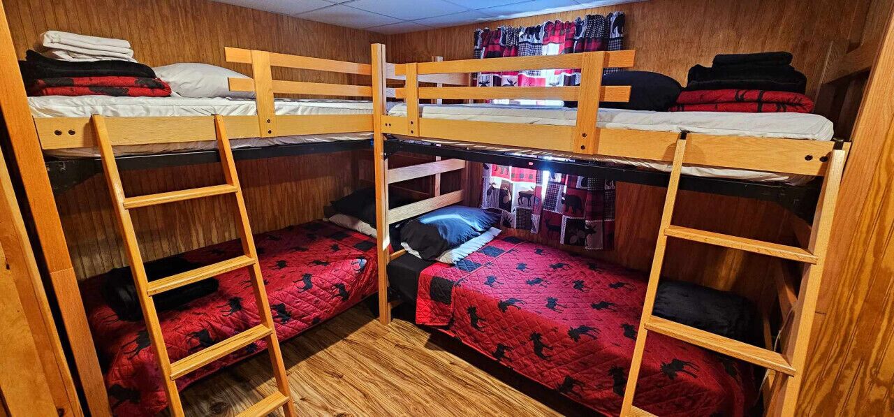 Ein Mehrbettzimmer mit Holzbettstapeln, einer Stehleiter und einer roten, gemusterten Bettdecke.