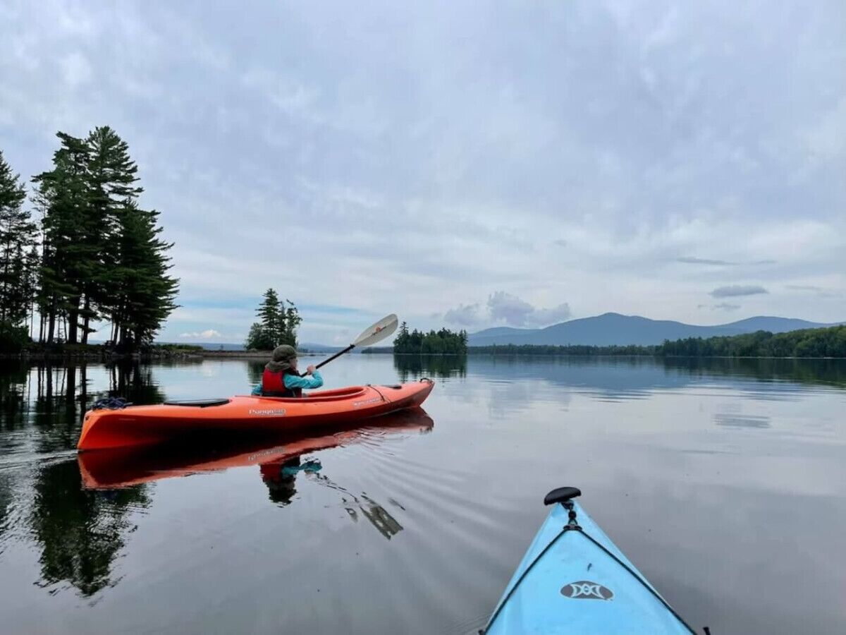 A kayaker in an orange kayak on a calm lake with a forest and mountains in the background.