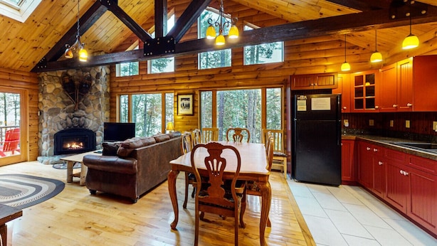 A spacious log cabin with a stone fireplace, wooden dining table, and red kitchen cabinets. - Loon Lodge on Wilson Pond (Greenville)