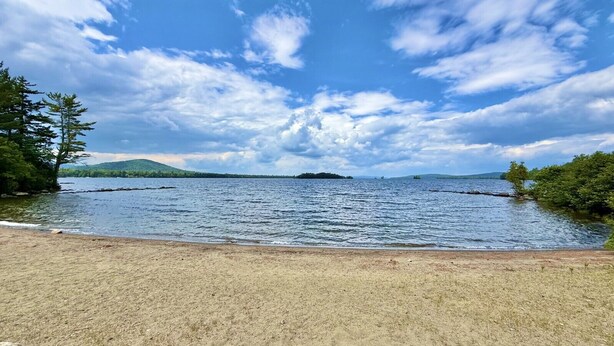 Blick auf einen See mit Sandstrand, Bäumen und sanften Hügeln im Hintergrund.