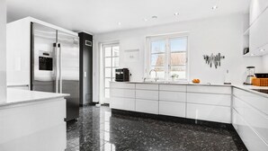 Une cuisine moderne avec des meubles blancs, un réfrigérateur en acier inoxydable, une machine à café et une fenêtre donnant sur une maison.