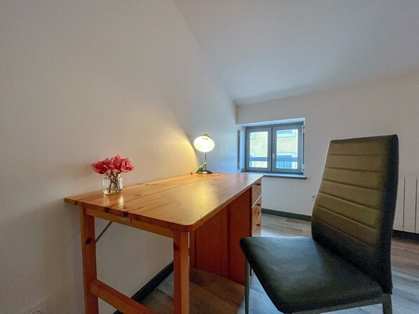 A room with a wooden desk, a chair, a small table with a vase of flowers, a lamp, and a window with a view of buildings.