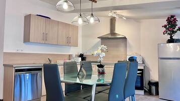 A modern kitchen with a dining area, featuring a glass table, metal chairs, a built-in oven, and pendant lighting.