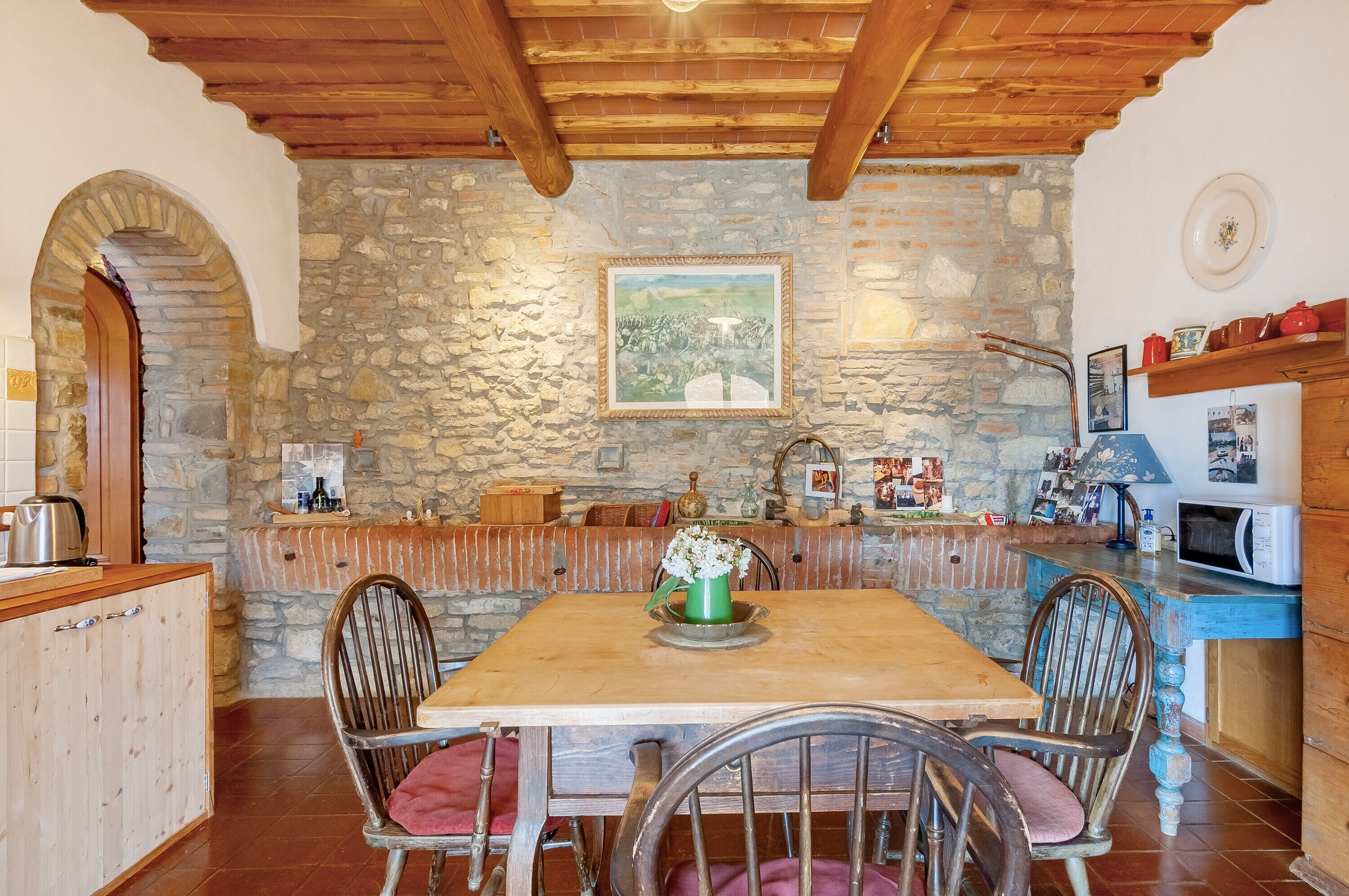 A rustic kitchen with stone walls, wooden beams, a dining table with chairs, and a stone fireplace.