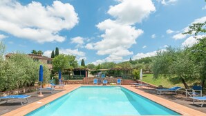Ein rechteckiger Swimmingpool mit blauen Liegestühlen und Sonnenschirmen, umgeben von viel Grün und blauem Himmel.