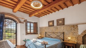 Ein Schlafzimmer mit Steinwand, Holzbalkendecke, einem Bett mit gemustertem Überwurf, einem Nachttisch mit Lampe und Blick durch ein Fenster.