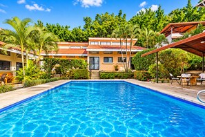 A swimming pool with a clear blue water surface, surrounded by lush greenery and a multi-story building with a red-tiled roof.