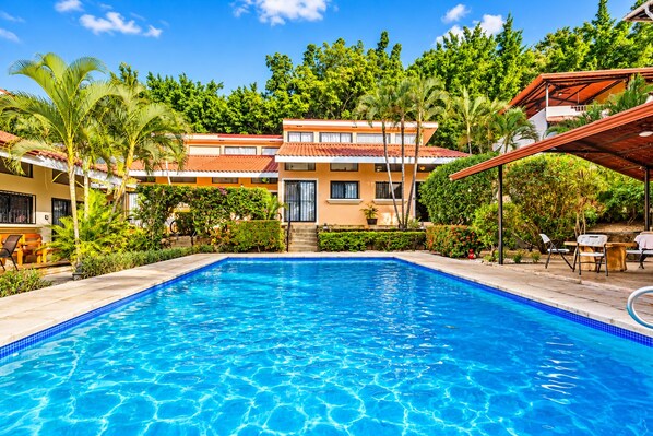 A swimming pool with a clear blue water surface, surrounded by lush greenery and a multi-story building with a red-tiled roof.