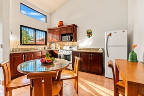 A kitchen with wooden cabinets, a stove, a microwave, and a refrigerator. There is a dining table with chairs and a fruit bowl on it.