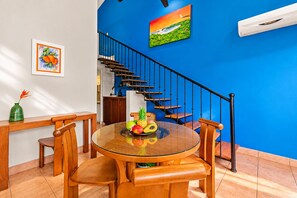 A dining area with a round glass table, wooden chairs, a fruit bowl, and a staircase with blue walls.