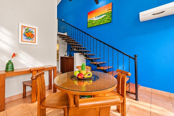 A dining area with a round glass table, wooden chairs, a fruit bowl, and a staircase with blue walls.
