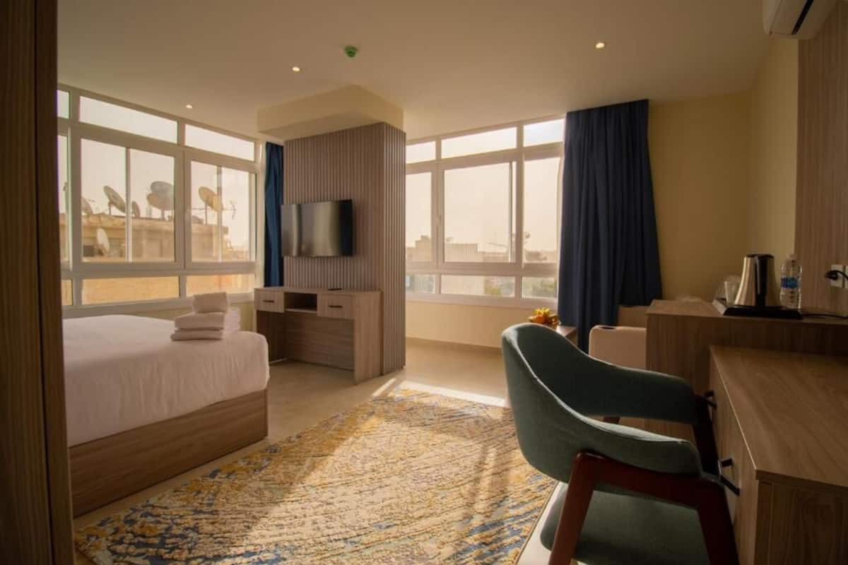 A hotel room with a bed, a desk, a chair, a TV, and large windows.